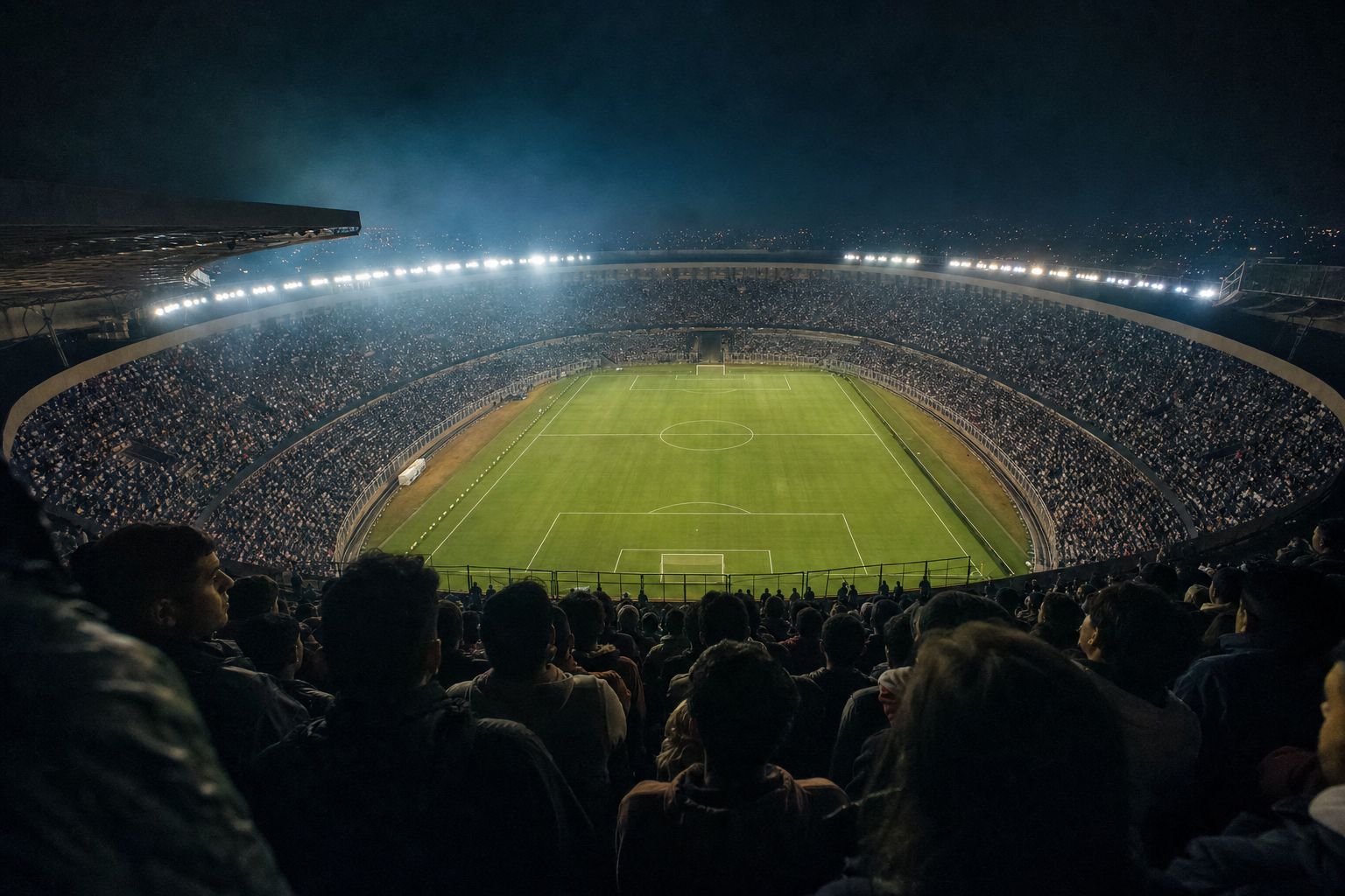Estadio iluminado de noche visto desde la bandeja alta, con la hinchada en primer plano