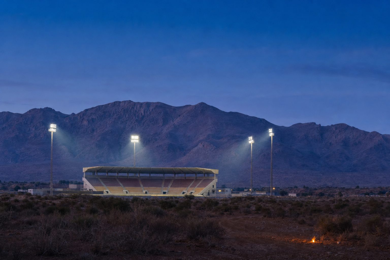 Estadio de fútbol iluminado al anochecer con una sierra de fondo, visto desde el descampado